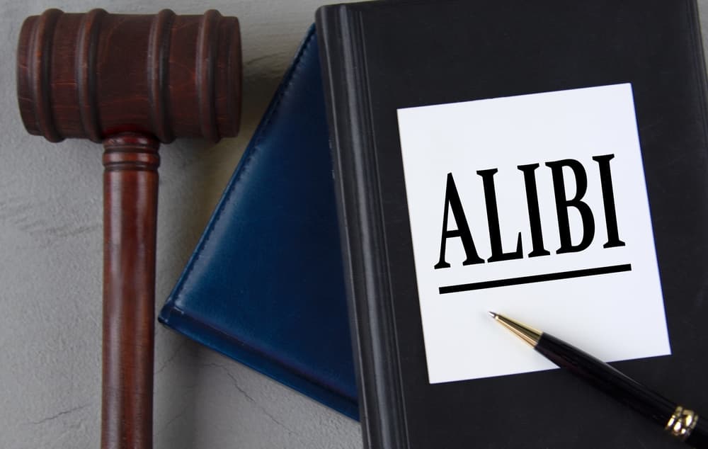 The word "ALIBI" written on a white sheet, placed alongside leather notebooks, a judge's gavel, and a pen.