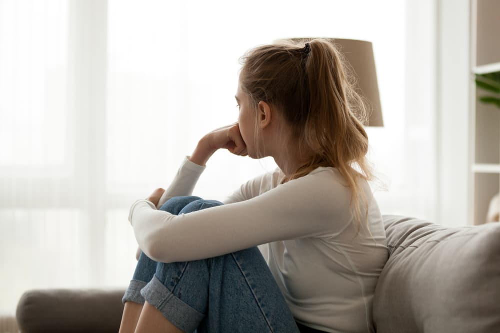 Young woman sitting on a couch, looking away from the window, appearing frustrated and confused about personal issues.