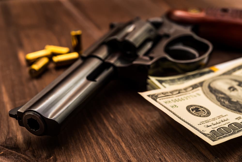 A revolver on a wooden surface beside $100 bills and bullets, symbolizing a robbery scenario.