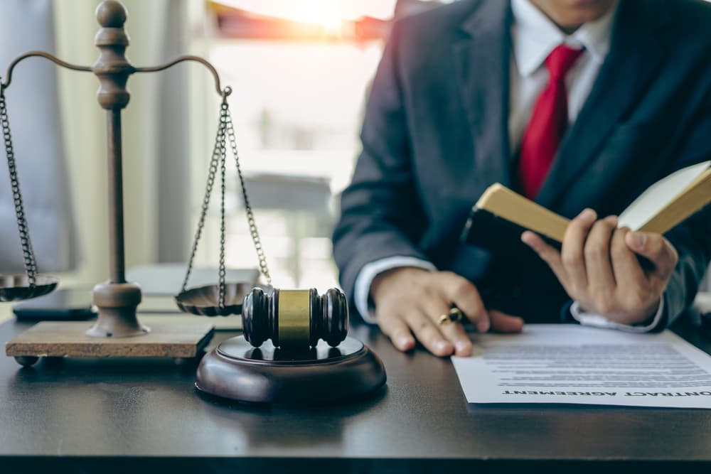 A lawyer in a suit working at a wooden table in an office, with documents, a laptop, a judge's gavel, and a scale of justice nearby. This close-up scene depicts legal advice and services.