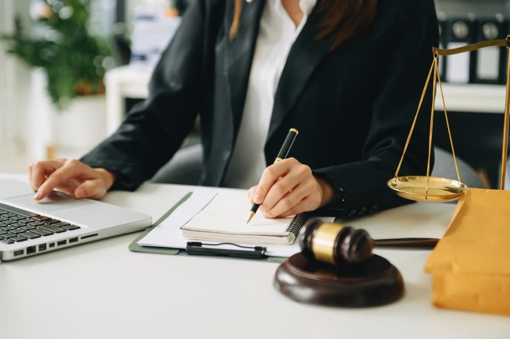 Justice and law concept: An Asian woman lawyer working in a courtroom, with a judge's gavel nearby, using a laptop and digital tablet on a white desk.